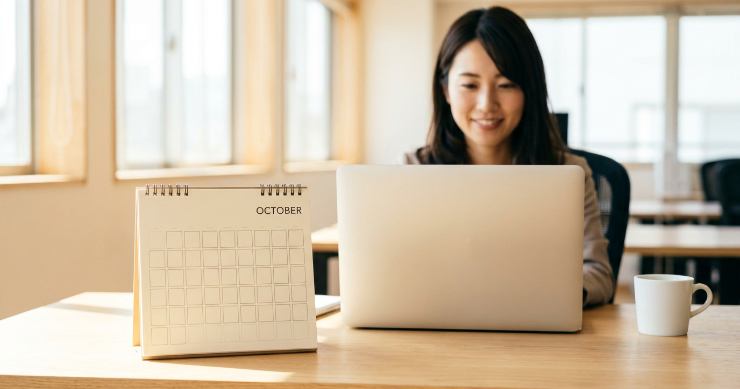 ノートパソコンの横に置かれた使いやすそうな卓上カレンダーと、背景で作業する日本の女性社員の写真
